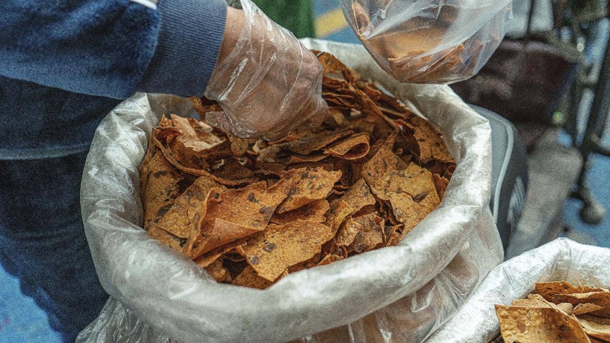 Seseorang mengenakan sarung tangan plastik sedang mengambil jajanan kering dari wadah besar berisi keripik berwarna cokelat.