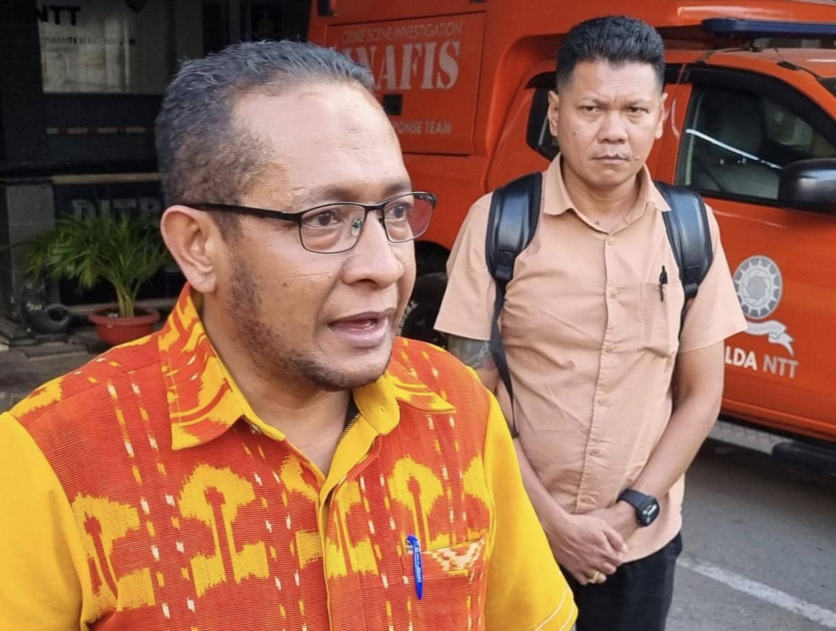 Seorang pria berbicara di depan gedung dengan latar belakang mobil oranye bertuliskan Polda NTT dan seorang pria lain berdiri di belakangnya.