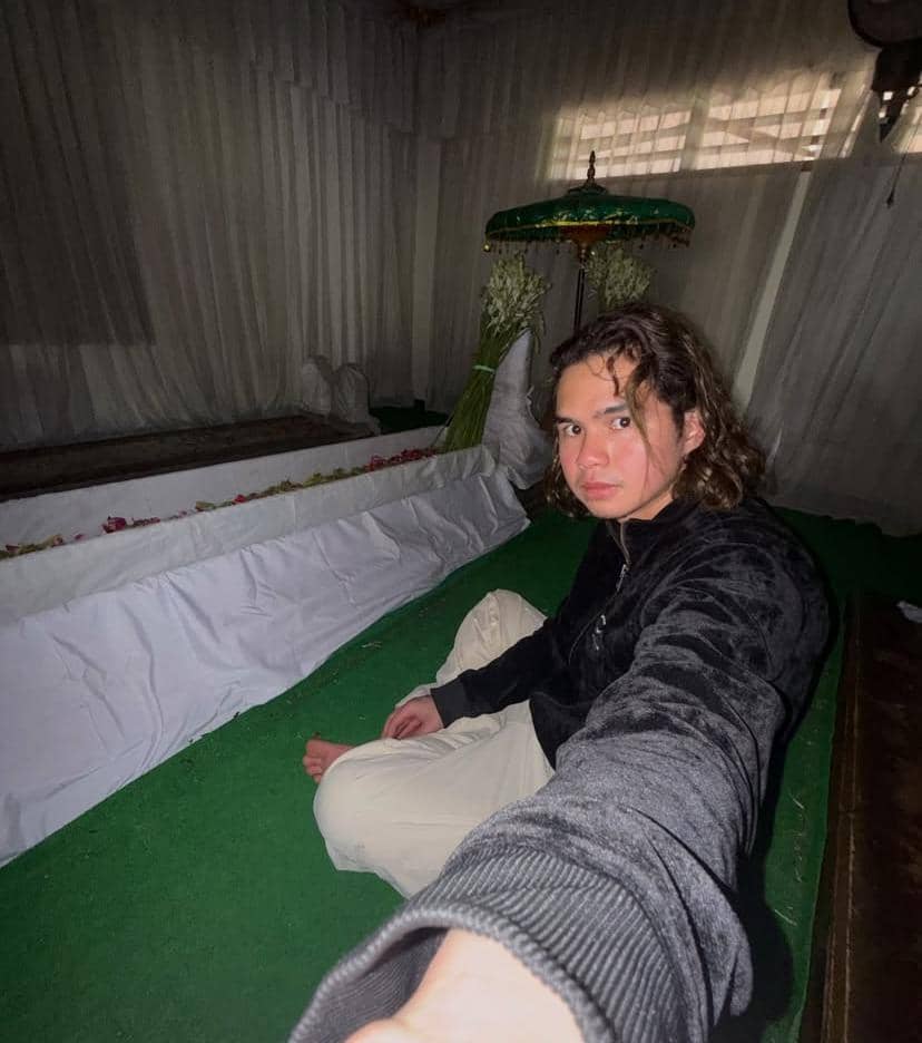 Seorang pria berambut panjang mengenakan jaket hitam duduk bersila di area makam yang ditutupi kain putih saat wisata religi di Tuban.