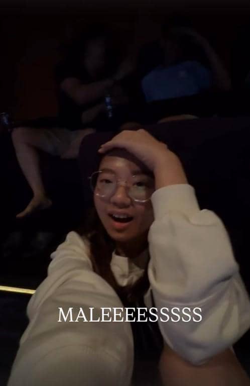 Seorang perempuan berkacamata berekspresi kaget sambil memegang kepala di ruangan gelap dengan tulisan 'MALEEESSSSS' di layar.