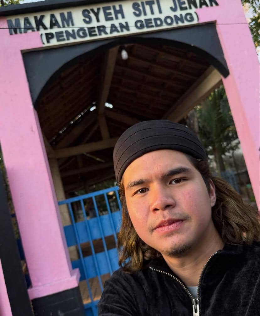 Seorang pria berpose di depan gerbang bertuliskan Makam Syekh Siti Jenar di Tuban dengan latar bangunan bercat merah muda dan hitam.