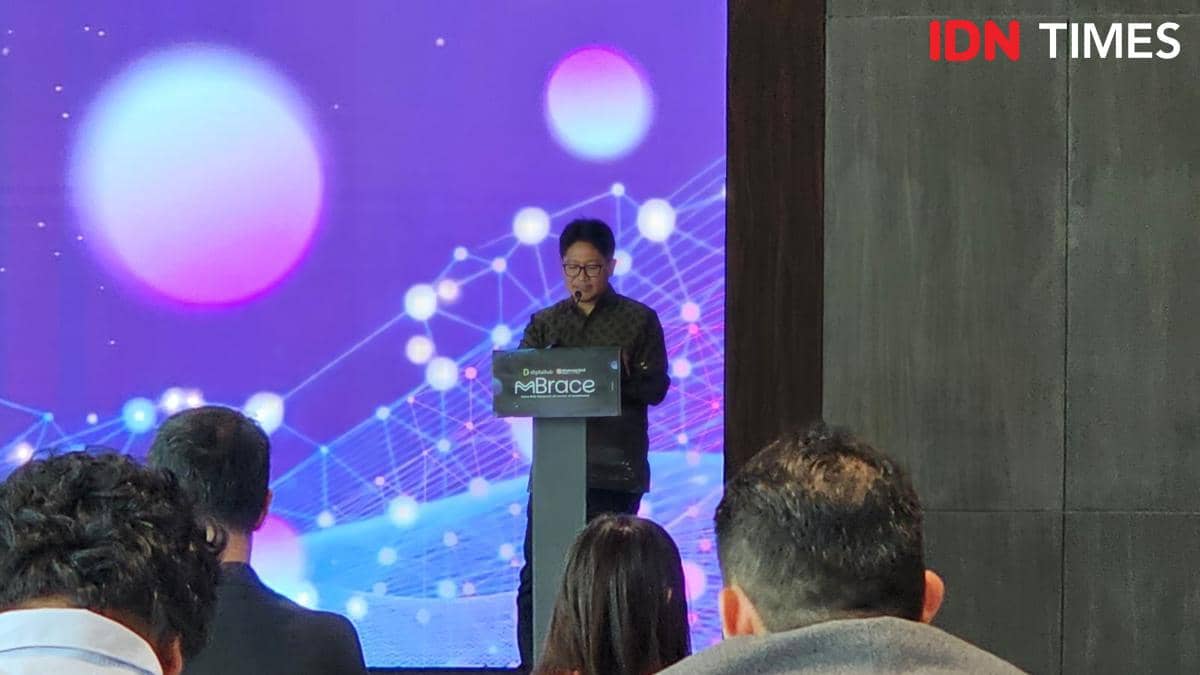 Seorang pembicara berdiri di podium dengan latar belakang visual digital berwarna ungu dan biru di depan audiens dalam sebuah acara.