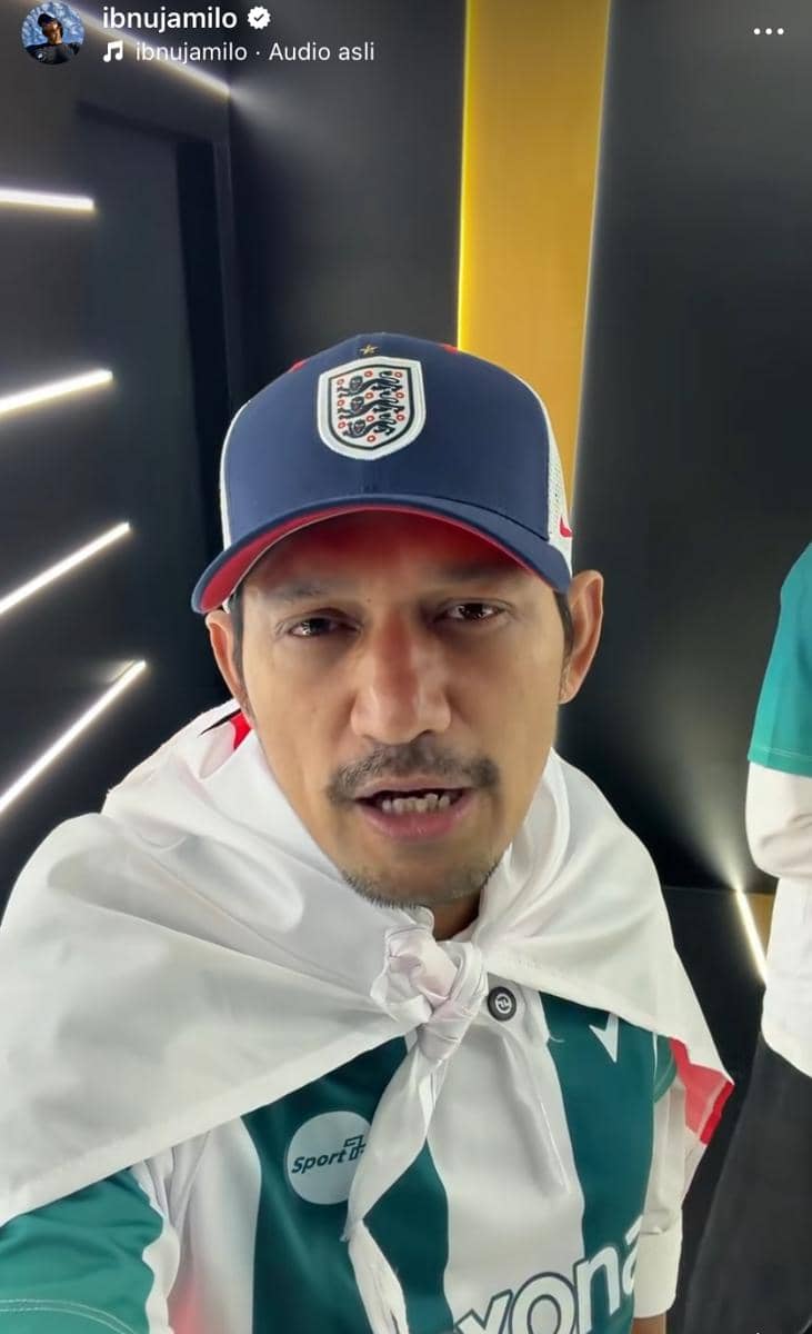 Seorang pria mengenakan topi biru bergambar logo tim sepak bola dan berselimut kain putih sambil mengenakan jersey hijau putih.