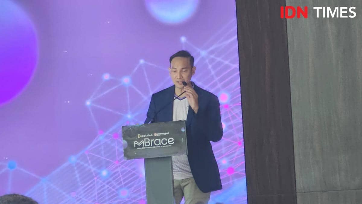 Seorang pria berbicara di atas panggung dengan latar belakang ungu bertema teknologi dan podium bertuliskan MBrace.