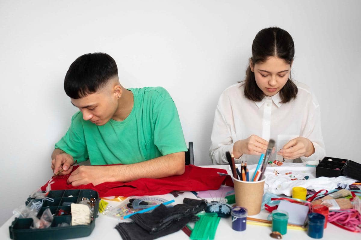 Dua orang sedang membuat kerajinan tangan di meja dengan berbagai bahan seperti kain, benang, cat, dan peralatan seni.