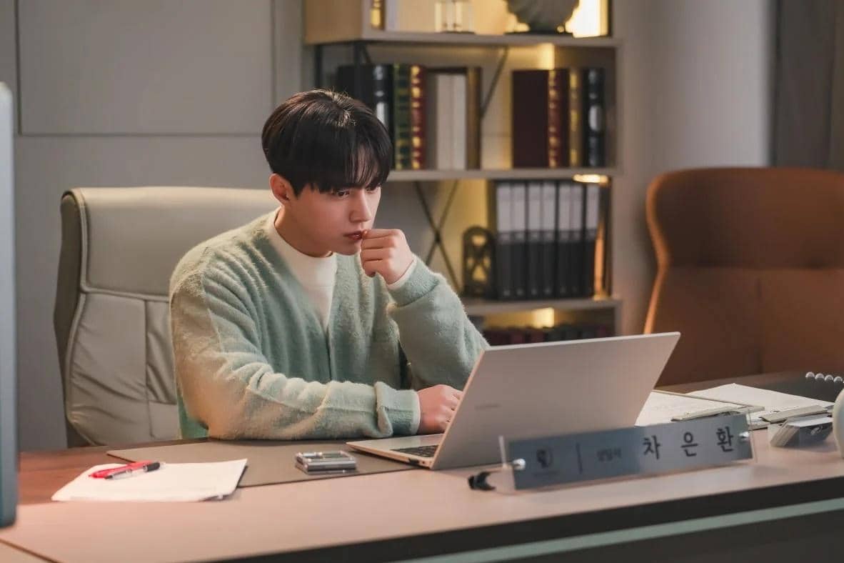 Seorang pria duduk di meja kerja dengan laptop di depannya, tampak serius menatap layar di ruang kantor modern dengan rak buku di belakangnya.