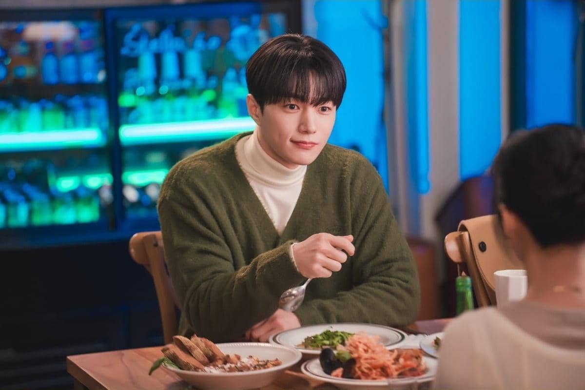 Seorang pria mengenakan sweater hijau duduk di meja makan dengan beberapa hidangan di depannya dalam adegan drama Love In Sync.