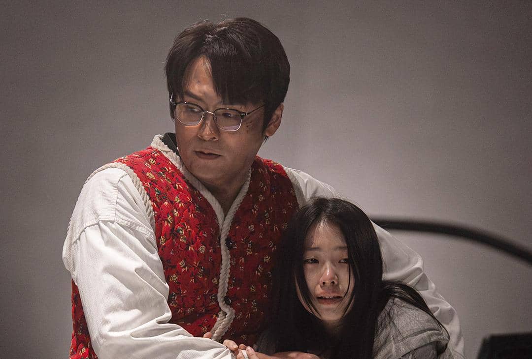 Seorang pria berkacamata mengenakan rompi merah bermotif bunga memeluk seorang wanita yang tampak ketakutan dalam adegan drama Korea Agent Kim Reactivated.