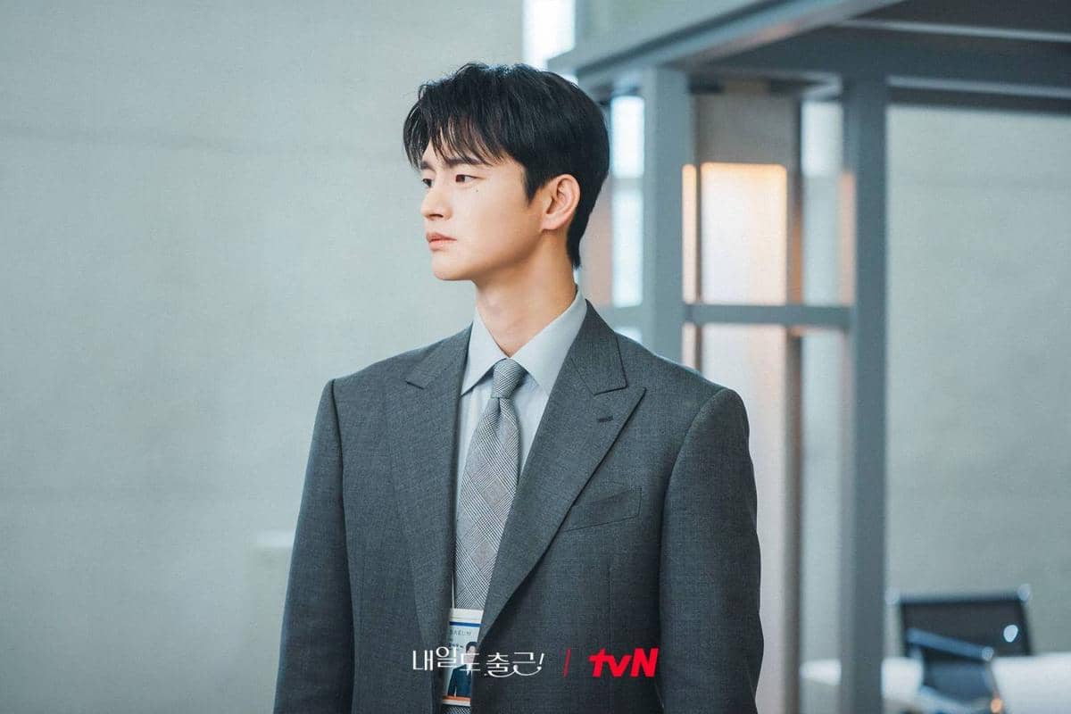 Seorang pria mengenakan setelan jas abu-abu dan dasi berdiri di dalam ruangan kantor dalam cuplikan drama Korea See You at Work Tomorrow.