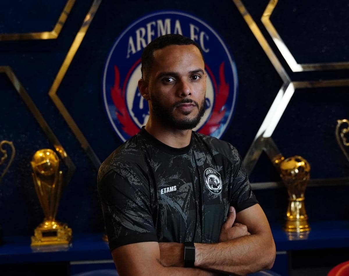 Seorang pemain Arema FC berpose dengan tangan terlipat di depan logo klub dan beberapa trofi emas di latar belakang.