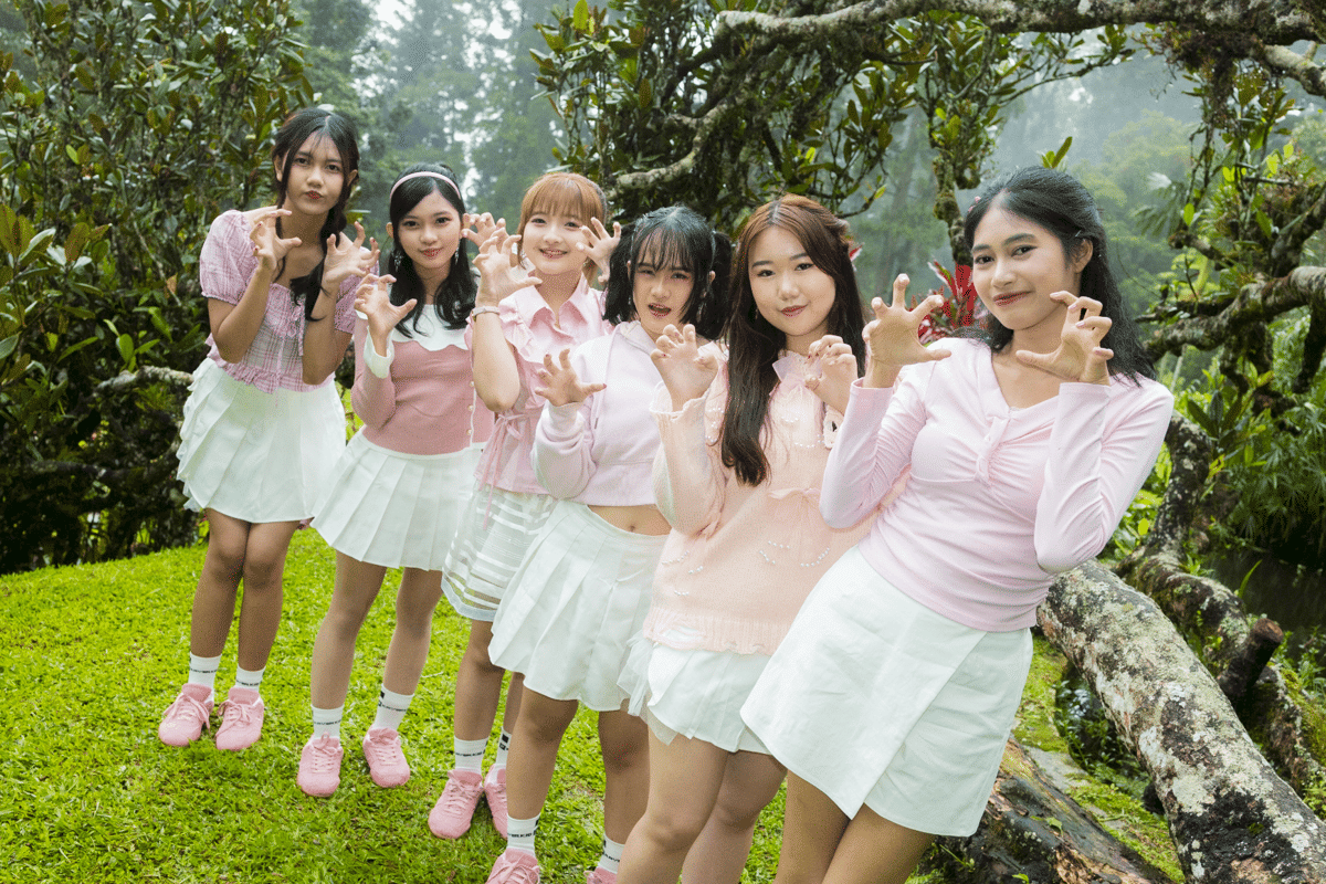 Enam anggota grup idola perempuan berpose dengan gaya lucu mengenakan atasan berwarna merah muda dan rok putih di taman hijau.
