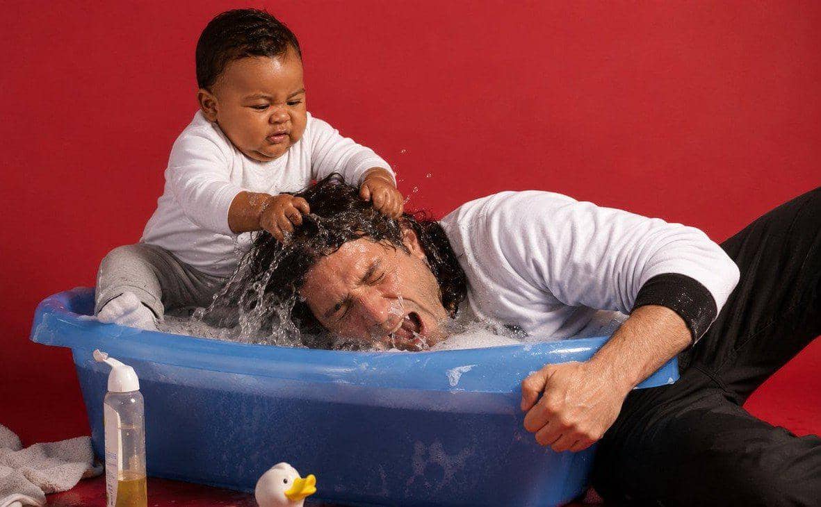Seorang bayi menarik rambut pria dewasa yang kepalanya masuk ke dalam bak mandi plastik biru dengan latar belakang merah.