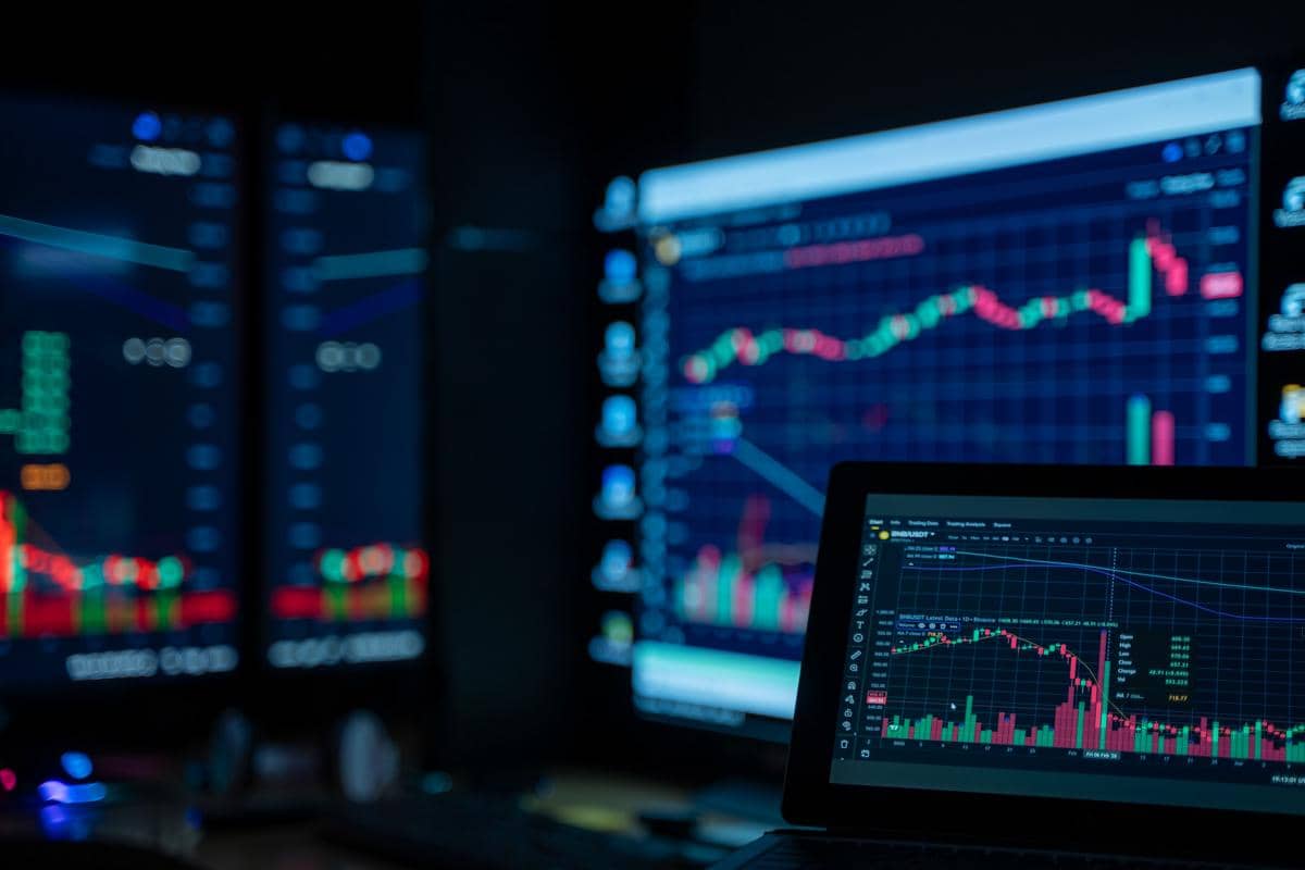 Beberapa layar komputer menampilkan grafik dan data pasar saham berwarna-warni sebagai ilustrasi diversifikasi aset investasi.