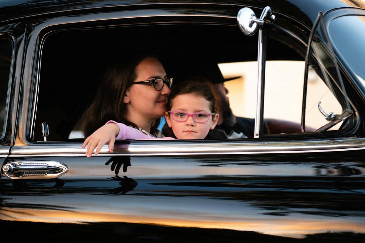 Anak kecil berkacamata duduk di dalam mobil hitam bersama orang dewasa, melihat keluar jendela dengan ekspresi tenang.