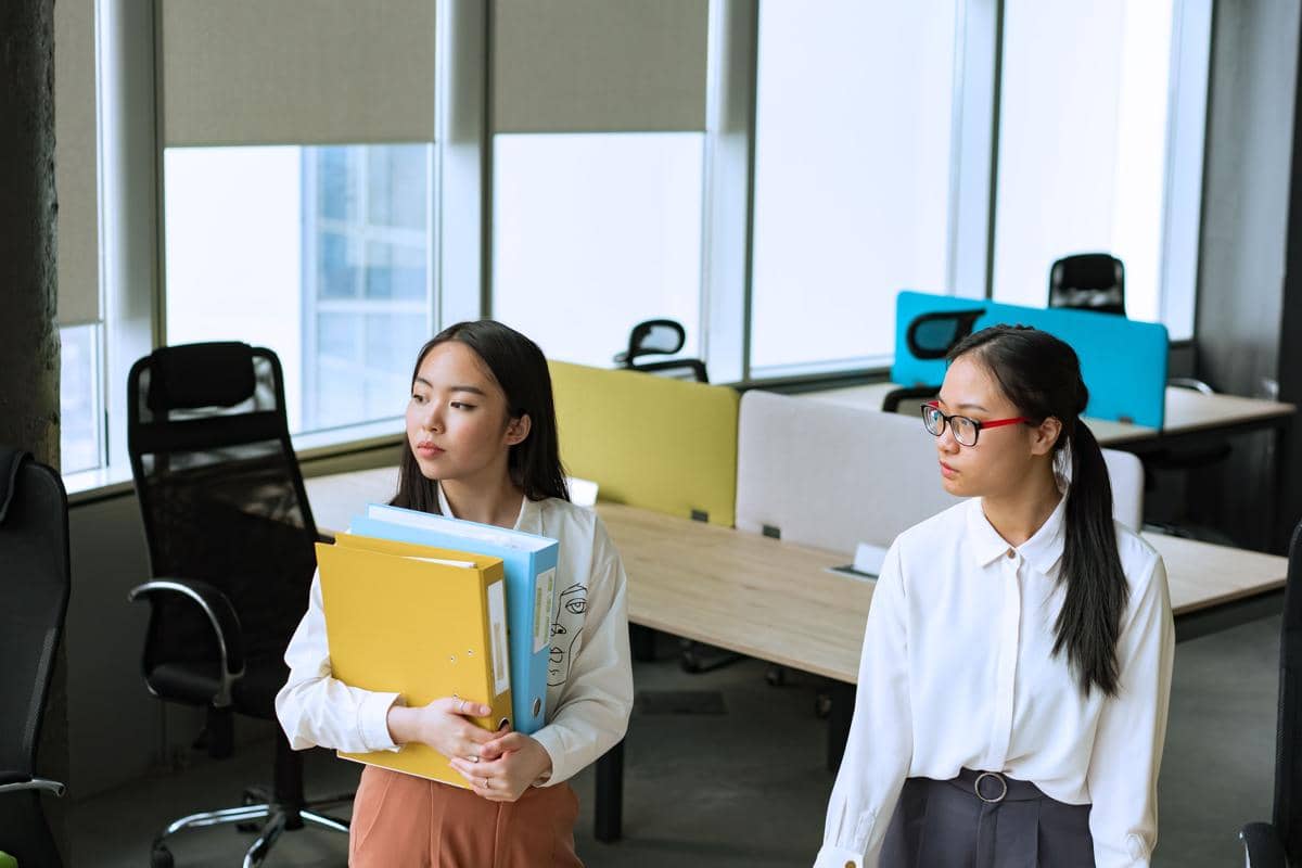 Dua perempuan di kantor, salah satunya memegang beberapa map dokumen berwarna kuning dan biru sambil berdiri di dekat meja kerja.