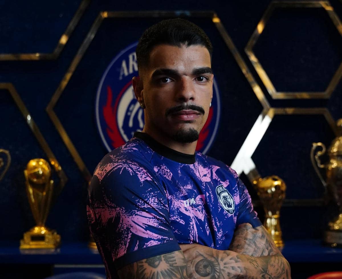 Seorang pemain Arema FC mengenakan jersey ungu bermotif berdiri dengan latar logo klub dan beberapa trofi emas di belakangnya.
