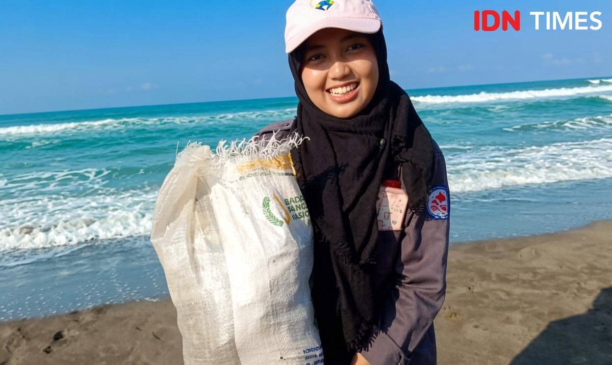 Perempuan berhijab tersenyum sambil memegang karung berisi sampah di tepi pantai dengan latar ombak laut biru yang tenang.
