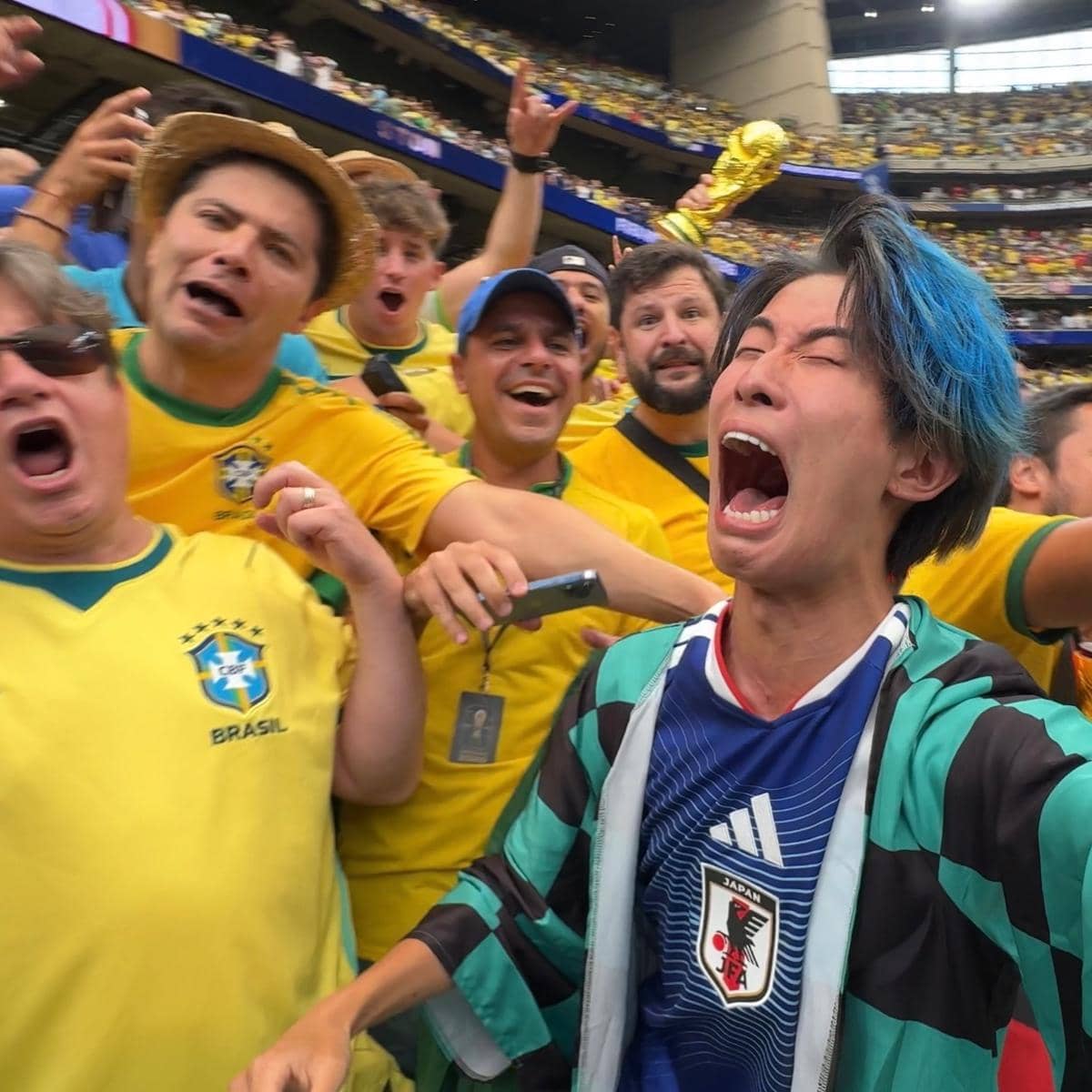 Seorang pria mengenakan jersey timnas Jepang berteriak di tengah kerumunan pendukung Brasil yang mengenakan seragam kuning di stadion sepak bola.