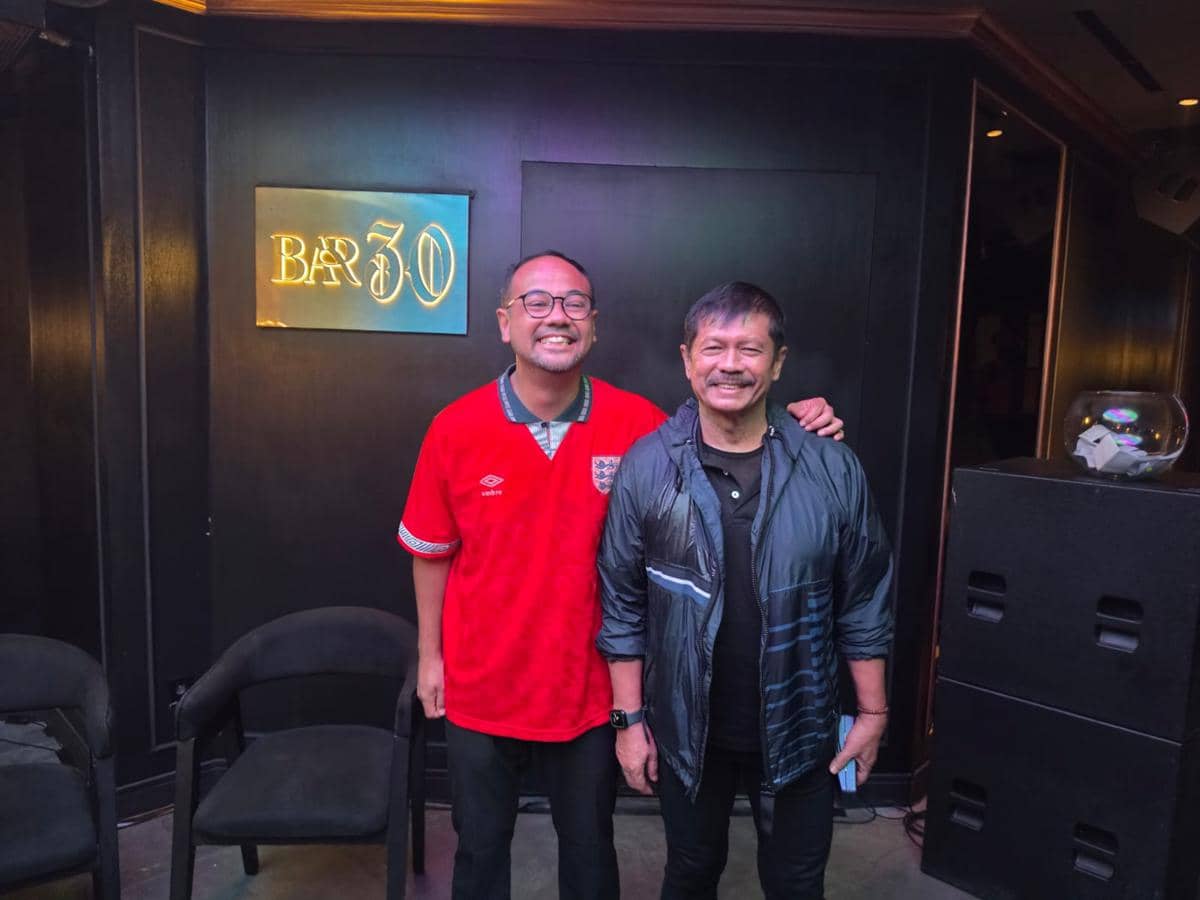 Dua pria berpose bersama di depan papan bertuliskan Bar 30, salah satunya mengenakan jaket hitam dan lainnya memakai jersey merah Inggris.