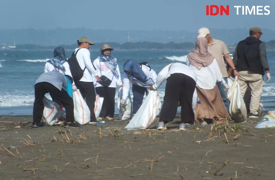 Sekelompok warga mengenakan pakaian kasual sedang memungut sampah plastik di tepi pantai Menganti sambil membawa karung besar.
