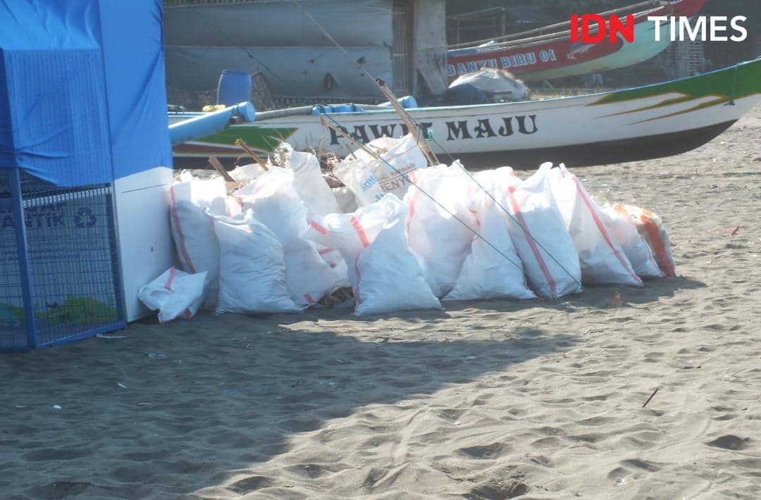 Tumpukan karung berisi sampah plastik di tepi pantai dekat dropbox biru dan perahu nelayan bertuliskan 'MAJU'.