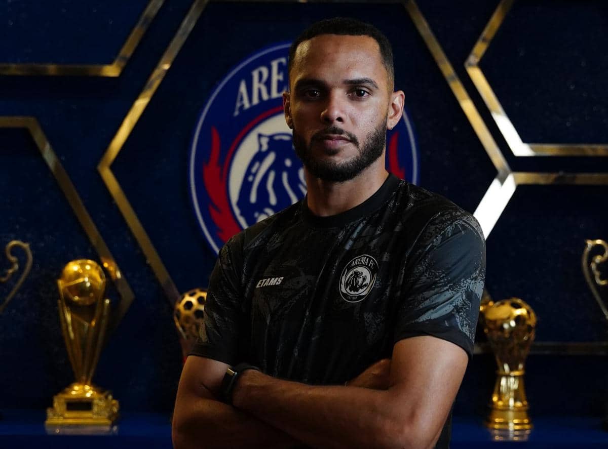 Seorang pemain Arema FC mengenakan jersey hitam berdiri dengan latar belakang logo klub dan beberapa trofi emas di belakangnya.