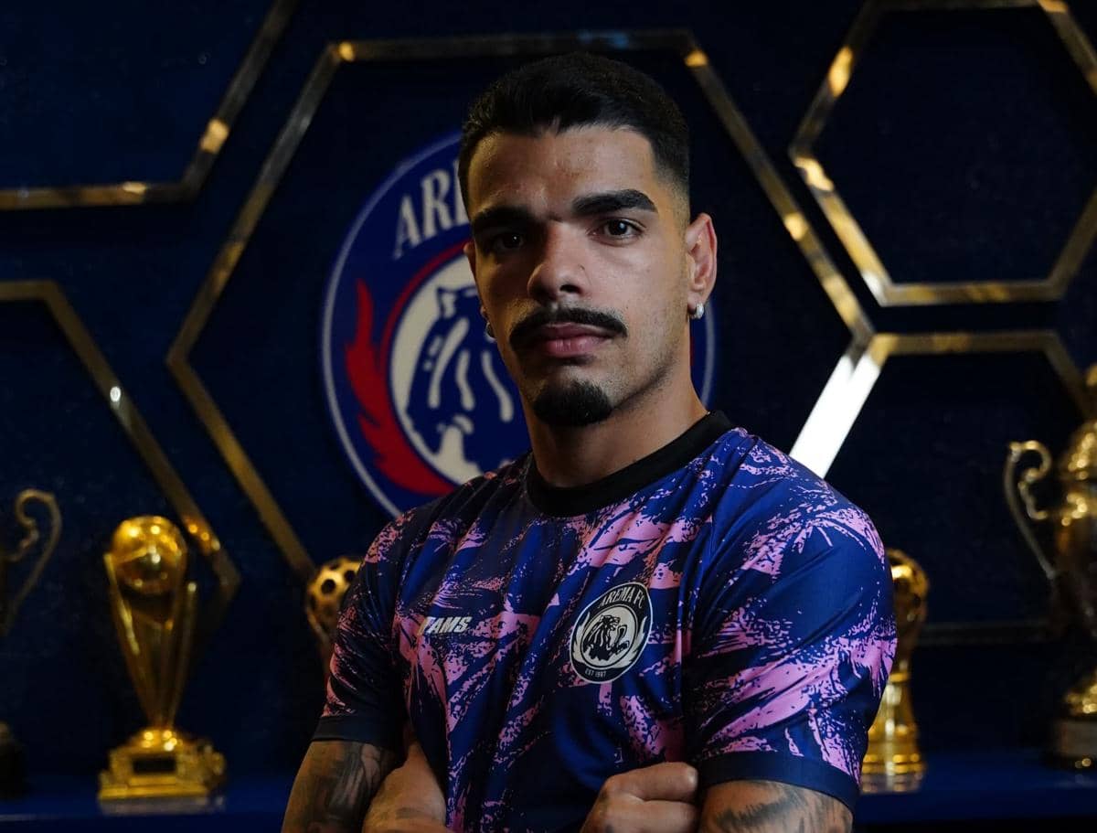 Seorang pemain Arema FC mengenakan jersey ungu dan biru berpose dengan latar belakang logo klub serta beberapa trofi emas.