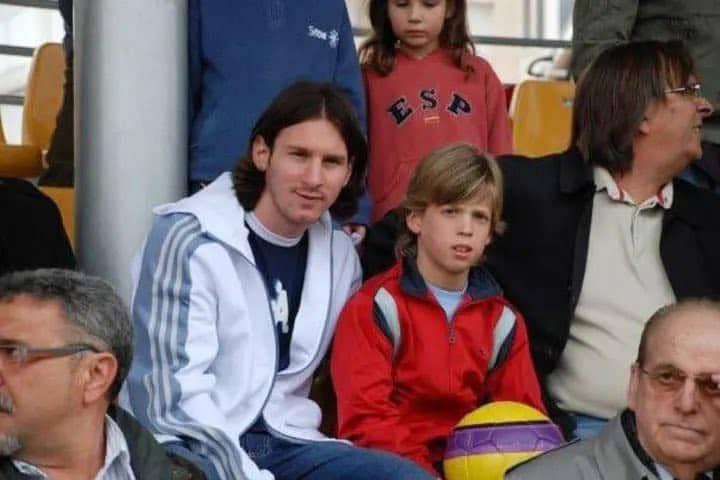 Seorang pria berjaket putih duduk di samping anak laki-laki berjaket merah yang memegang bola di antara penonton di tribun stadion.