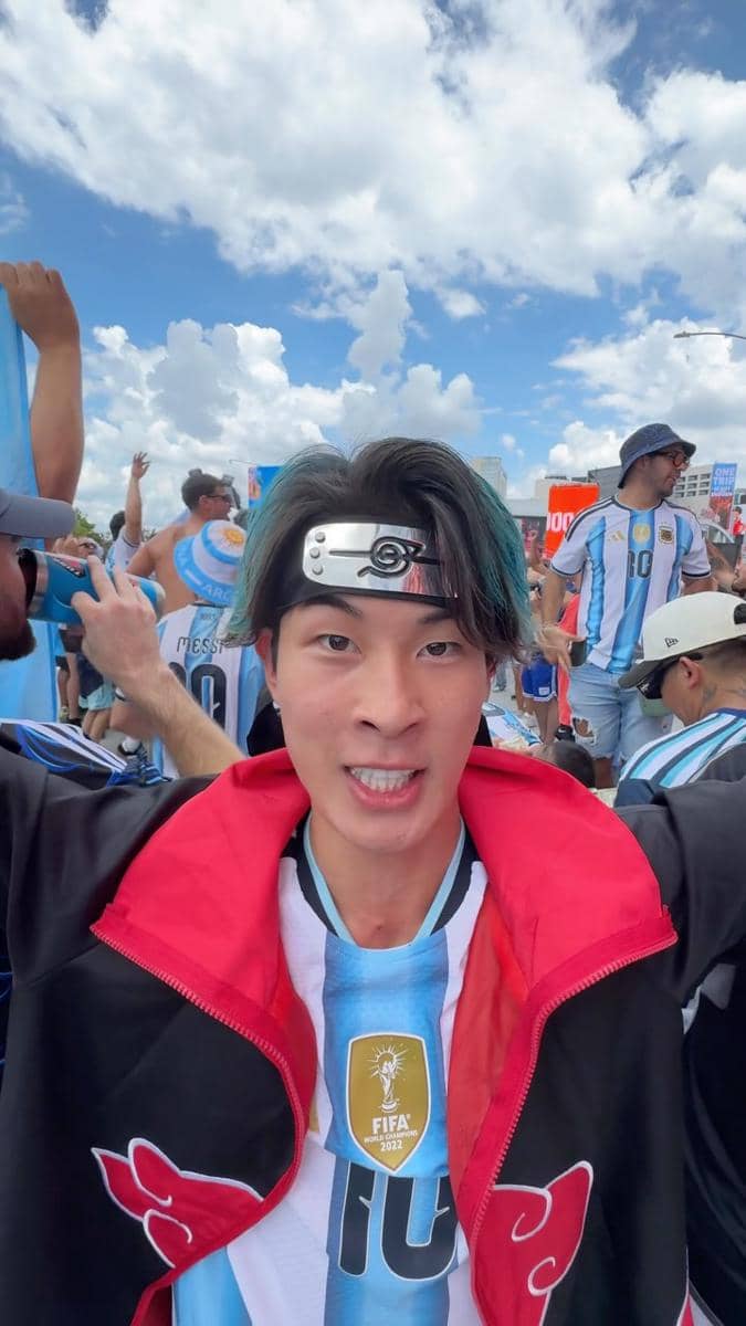 Seorang pria mengenakan jaket merah hitam dan ikat kepala anime, berpose di tengah kerumunan pendukung tim sepak bola Argentina.