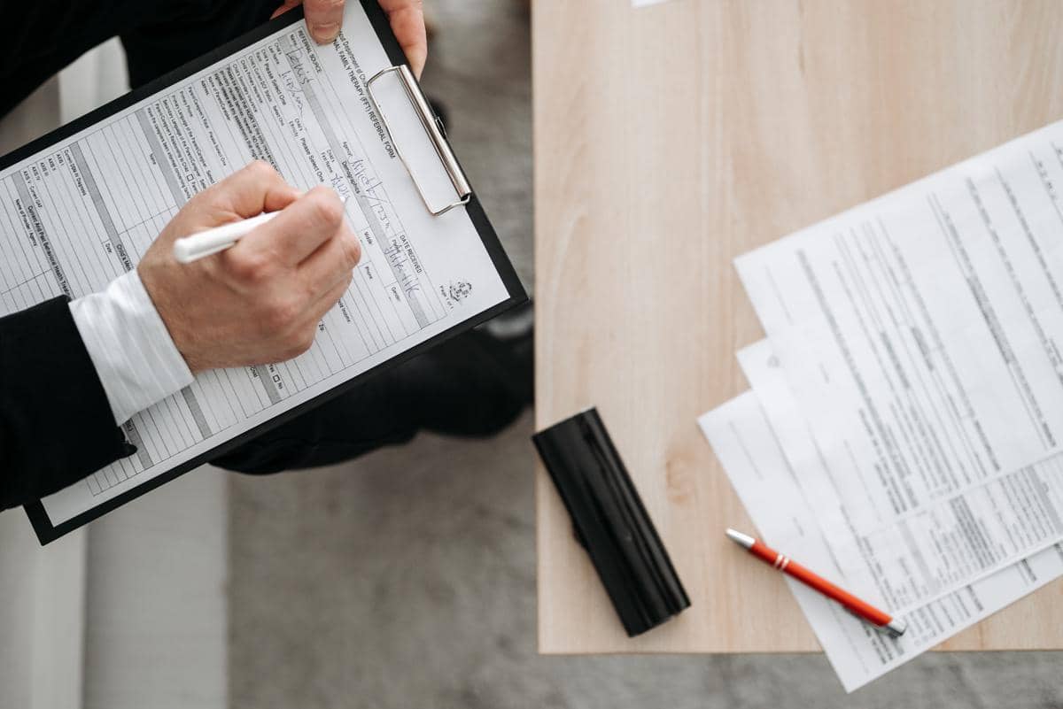 Seseorang mengenakan jas hitam sedang mengisi formulir lamaran kerja di atas clipboard dengan pena putih di meja kantor.