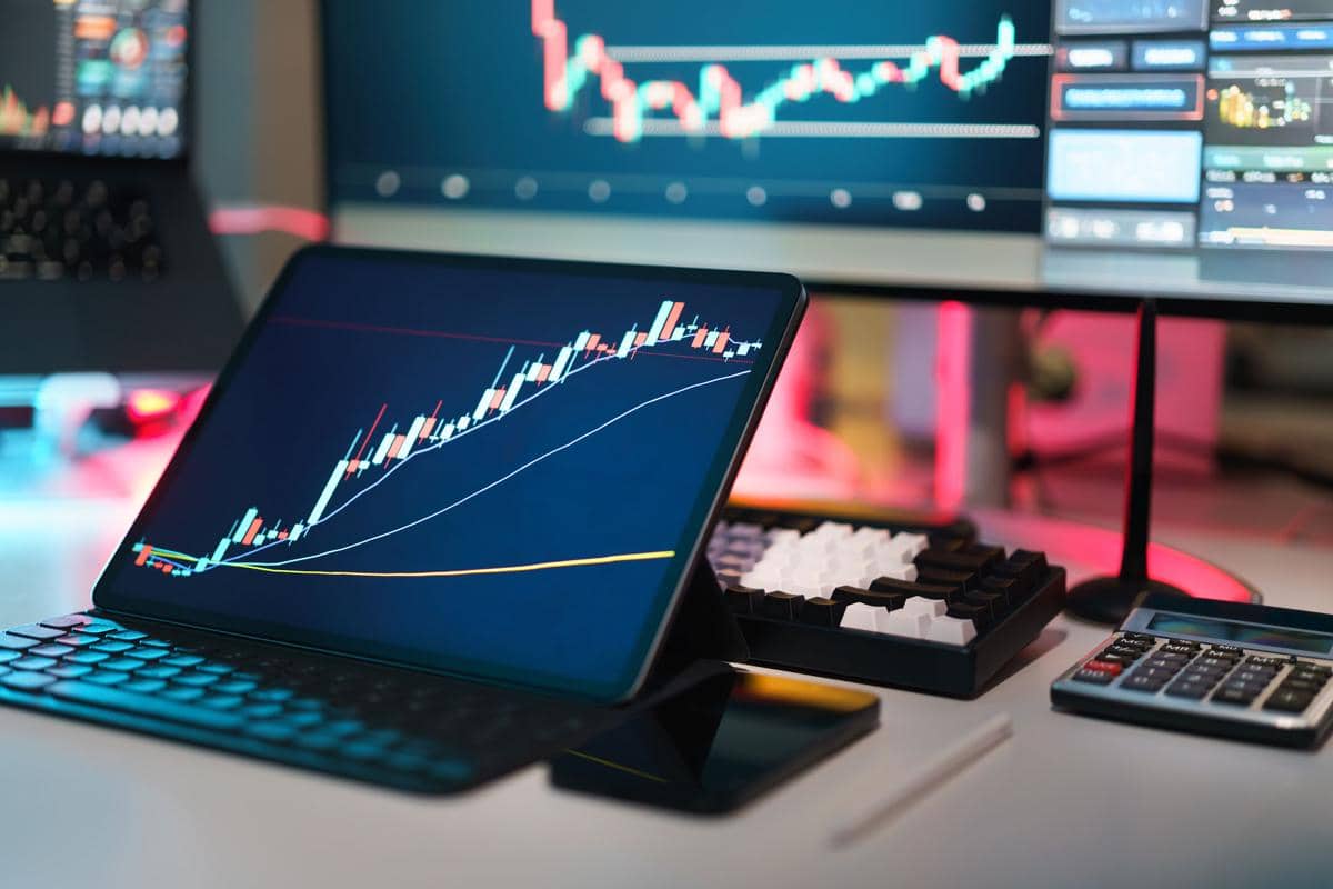 Grafik keuangan berwarna pada layar tablet di meja kerja dengan keyboard, monitor, dan kalkulator, menggambarkan analisis portofolio investasi.