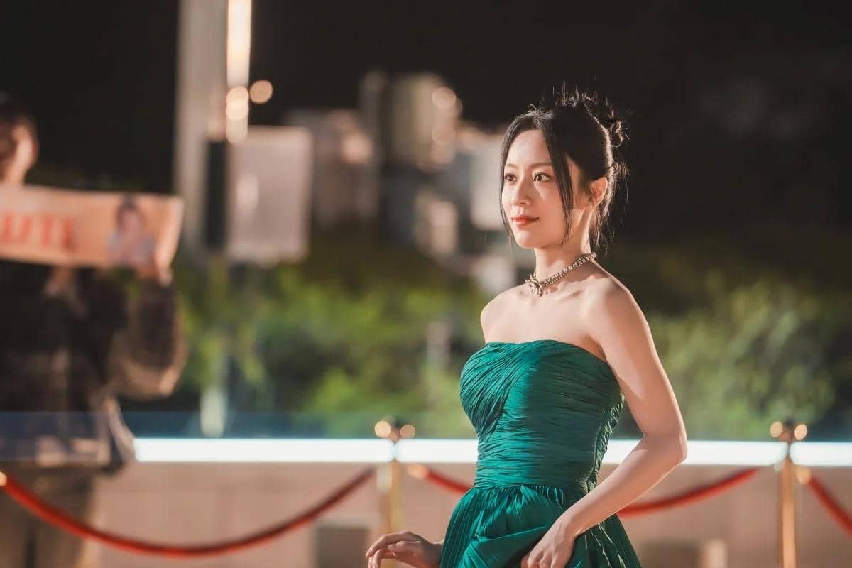 Seorang wanita mengenakan gaun hijau elegan tanpa tali dengan kalung mutiara, berdiri di area luar ruangan pada malam hari.