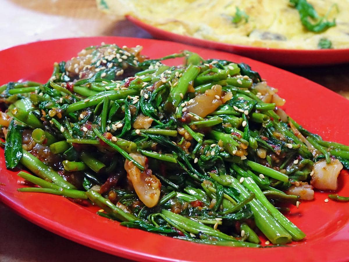 Tumis kangkung dengan potongan bawang dan taburan biji wijen disajikan di atas piring merah, tampak segar dan menggugah selera.