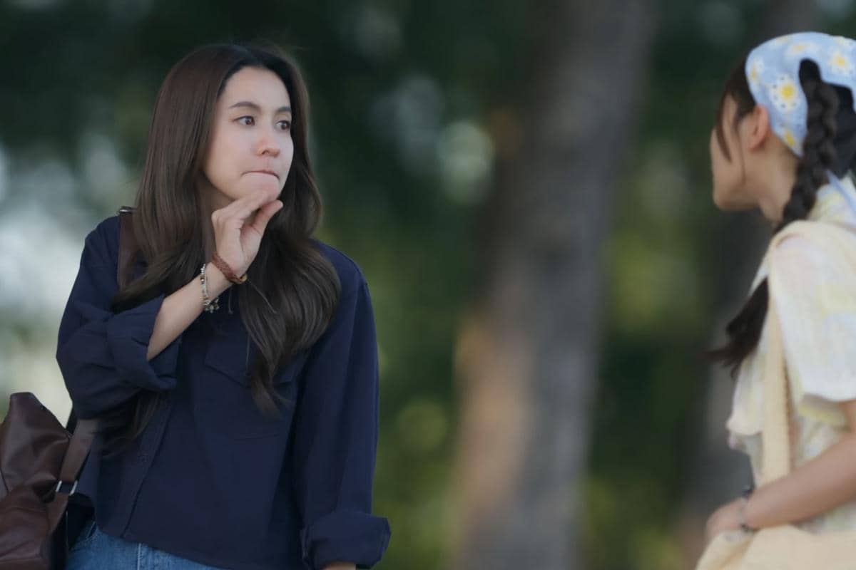 Dua perempuan sedang berbicara di luar ruangan, salah satunya mengenakan kemeja biru tua dan membawa tas cokelat, sementara yang lain mengenakan pakaian kuning muda.