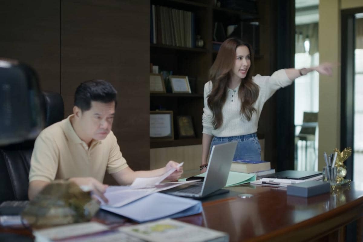 Seorang pria duduk di meja kerja dengan dokumen dan laptop, sementara seorang wanita berdiri di sampingnya sambil menunjuk dengan ekspresi tegas.