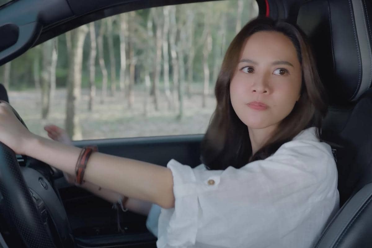 Seorang perempuan mengenakan kemeja putih duduk di kursi pengemudi mobil, memandang ke arah samping dengan latar pepohonan di luar jendela.
