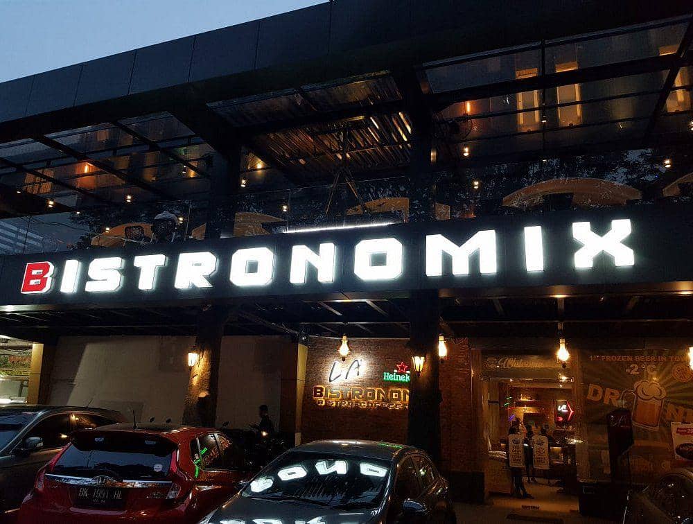 Tampak depan restoran Bistronomix pada malam hari dengan papan nama besar bercahaya dan beberapa mobil terparkir di depannya.