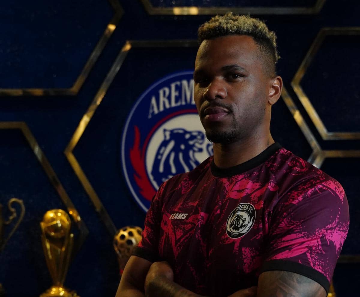 Pemain Arema FC mengenakan jersey berwarna merah marun dengan latar logo klub dan beberapa trofi emas di belakangnya.