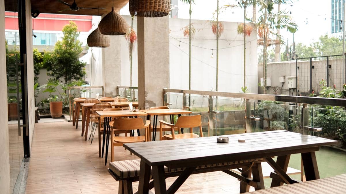 Area teras kafe dengan meja dan kursi kayu bergaya minimalis, dihiasi lampu gantung rotan dan tanaman hijau di sekelilingnya.