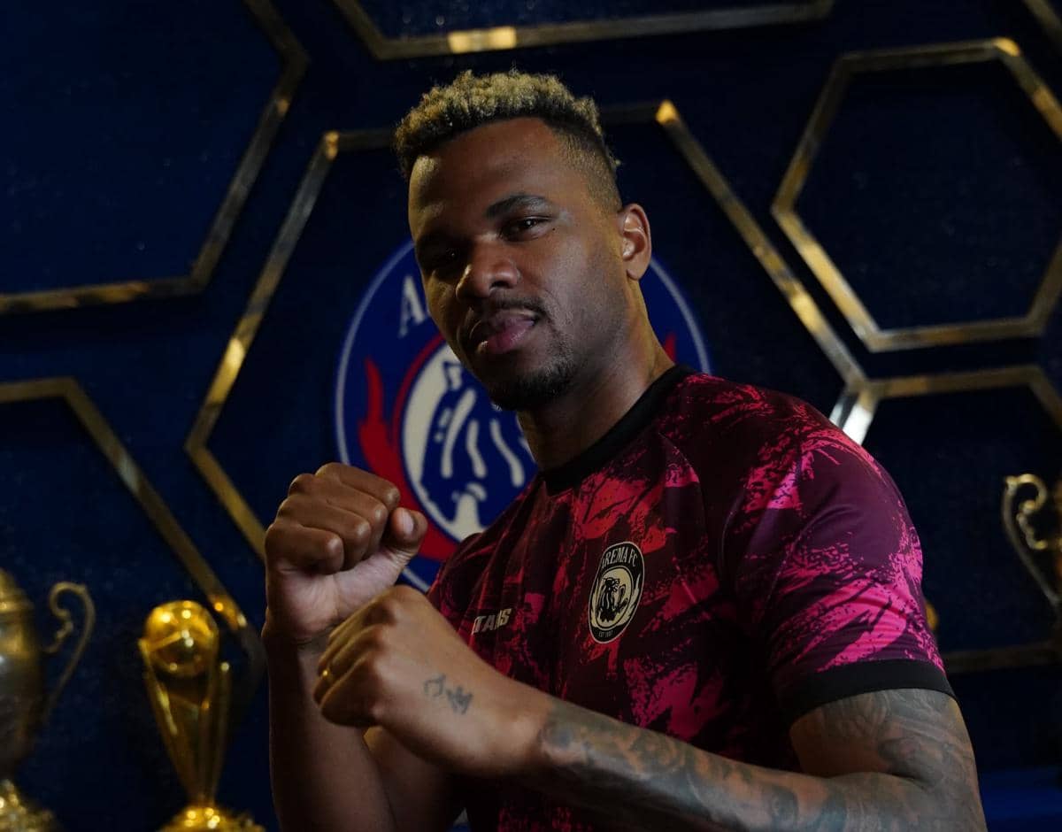 Seorang pemain Arema FC mengenakan jersey ungu berpose dengan tangan mengepal di depan logo klub dan piala di latar belakang.