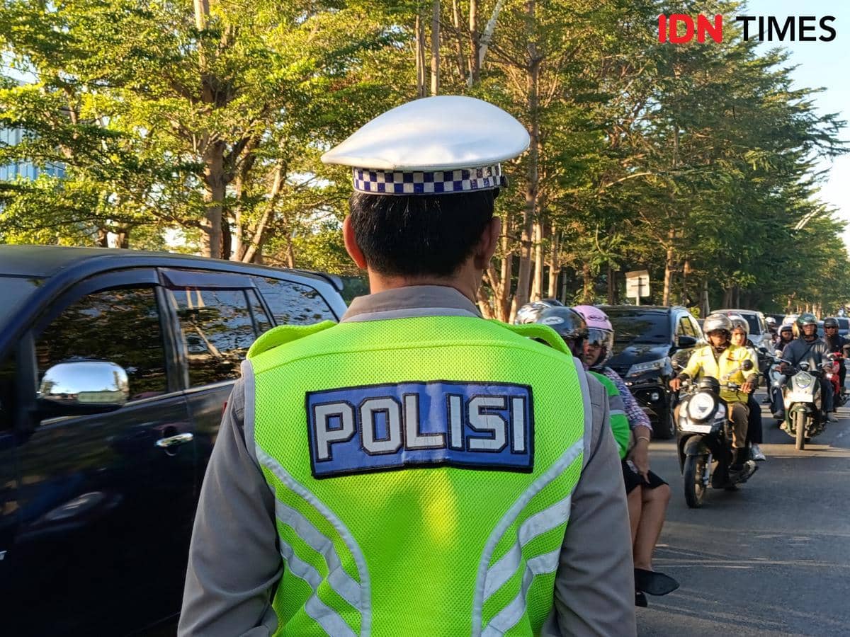 Seorang polisi lalu lintas mengenakan rompi hijau dan topi putih sedang mengatur kendaraan di jalan ramai Kota Kupang pada siang hari.