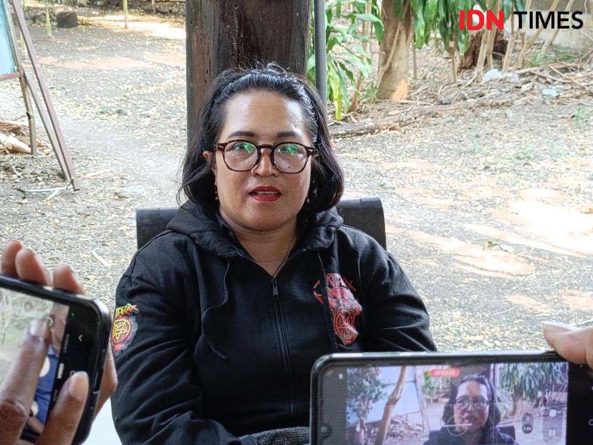 Seorang perempuan berjaket hitam dan berkacamata duduk di luar ruangan sambil berbicara, direkam menggunakan ponsel oleh beberapa orang.