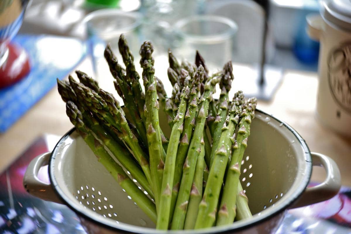 Beberapa batang asparagus segar berwarna hijau muda dengan ujung ungu disusun di dalam saringan logam di atas meja dapur.