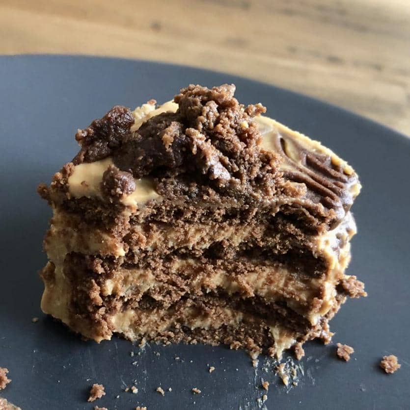 Potongan kue chocotorta berlapis biskuit cokelat dan krim karamel di atas piring hitam dengan tekstur lembut dan remah di sekitarnya.
