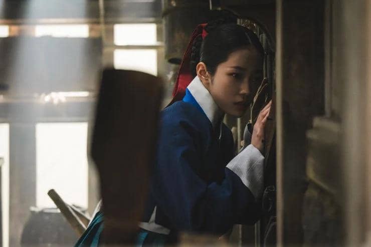 Seorang wanita mengenakan hanbok biru dengan pita merah di rambutnya tampak mengintip melalui celah di dinding kayu dalam suasana redup.