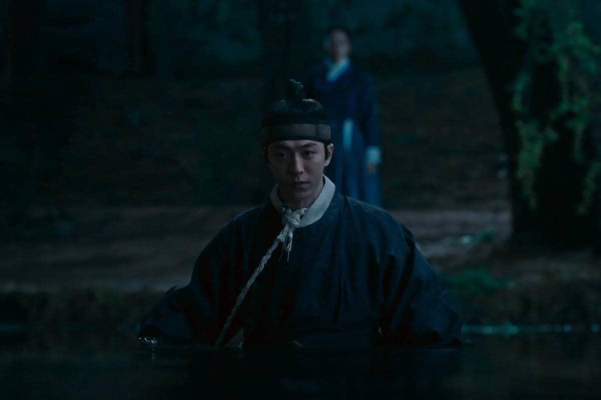 Seorang pria berpakaian tradisional berdiri di air pada malam hari dengan latar belakang hutan dalam adegan drama The East Palace.