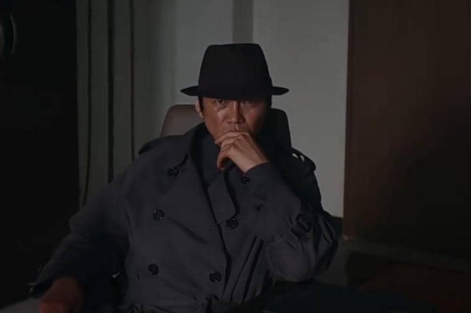 Seorang pria mengenakan mantel gelap dan topi hitam duduk di kursi dengan ekspresi serius dalam cuplikan drama Korea Agent Kim Reactivated.