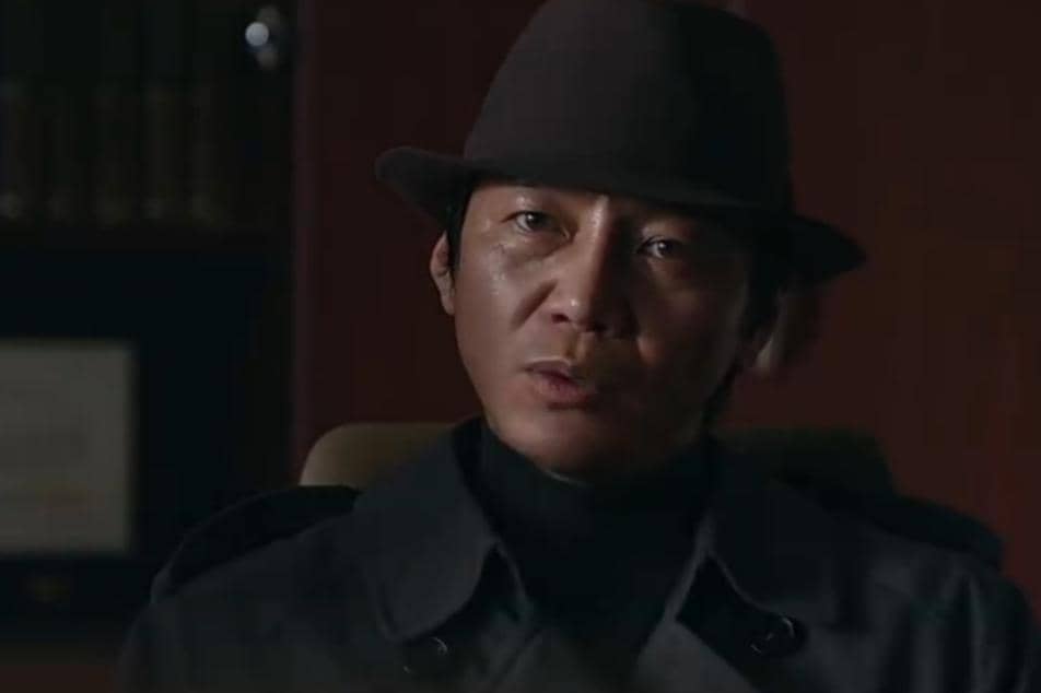 Seorang pria mengenakan mantel gelap dan topi fedora duduk di ruangan redup dalam cuplikan drama Korea Agent Kim Reactivated.