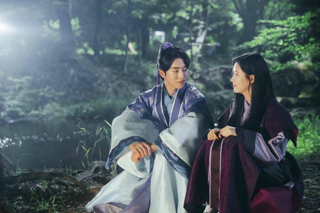 Dua karakter dalam drama Moon Lovers: Scarlet Heart Ryeo duduk berhadapan di tepi sungai pada malam hari dengan latar pepohonan hijau.