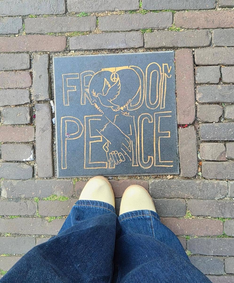 Sepasang kaki dengan sepatu krem berdiri di atas ubin bertuliskan 'Freedom Peace' dengan gambar burung merpati dan tangan berjabat.